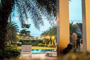 Zona junto a la piscina con un banco, palmeras y luces colgantes.
