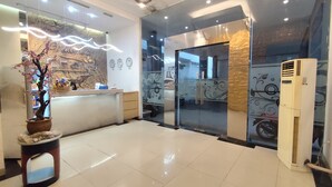 A hotel reception area with a reception desk, a potted plant, a wall clock, and a large air conditioning unit. - De La Charis Hotel (Tulungagung)