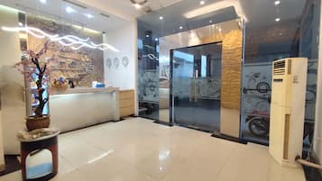 A hotel reception area with a reception desk, a potted plant, a wall clock, and a large air conditioning unit.