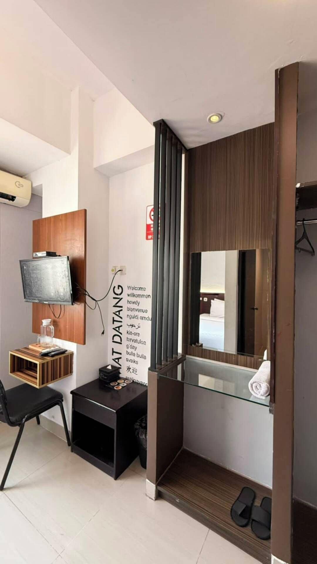Ein modernes Hotelzimmer mit einem an der Wand befestigten Flachbildfernseher, einem kleinen Holfnachtisch mit Fernbedienung, einem Glastischchen mit weißer Vase und einem Spiegel mit Holzrahmen.