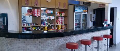 Un espace bar avec un comptoir carrelé, des étagères remplies de collations et trois tabourets de bar rouges.