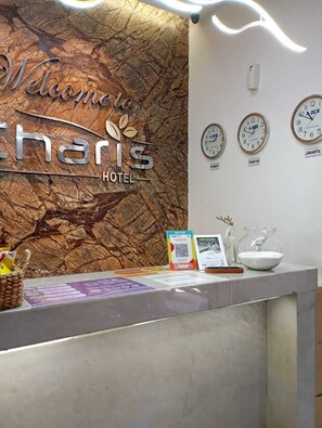 A reception desk with a 'Welcome to Charis Hotel' sign, clocks indicating time in Jakarta, Sydney, Tokyo, and a QR code for saving a room.