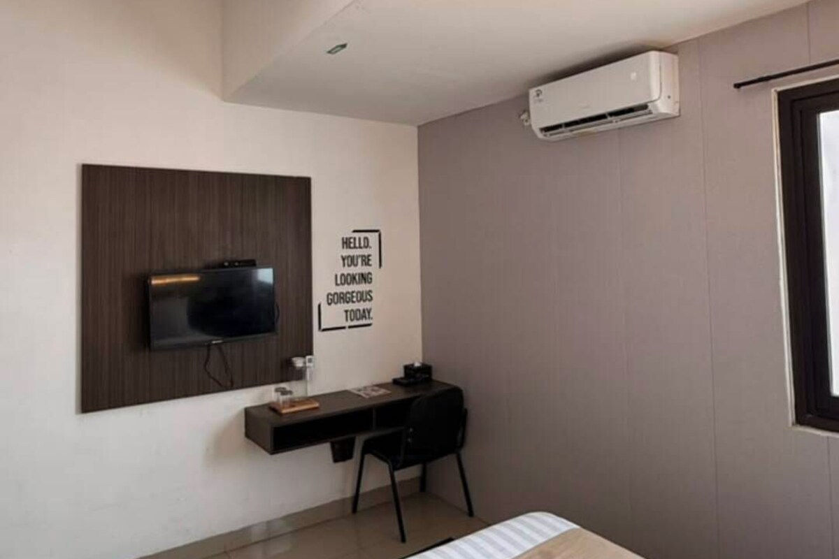A modern bedroom with a bed, a desk, a flat-screen TV, and an air conditioning unit.