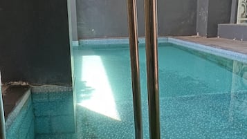 Ein Schwimmbecken mit einer Metallleiter, umgeben von einer dunklen Wand und einer Rasenfläche.