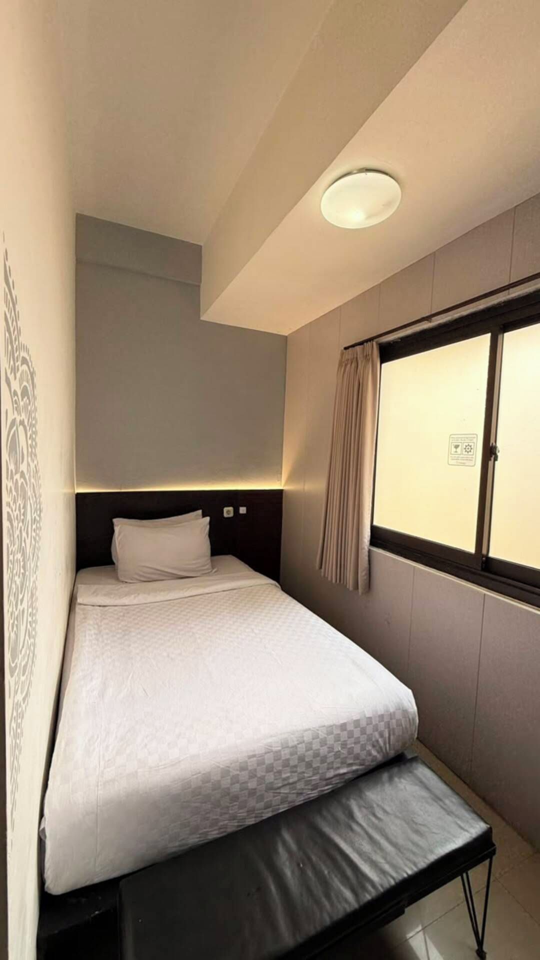 A single bed with a headboard, a window with curtains, and a wall with a decorative pattern.
