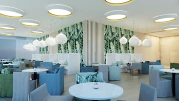 Ein modernes Restaurant mit runden Tischen, blau-grauen Sitzgelegenheiten und einer Wand mit tropischen Blattmotiven.