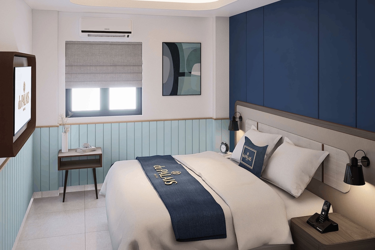 A hotel room with a bed, bedside table, lamp, and a wall-mounted air conditioner.