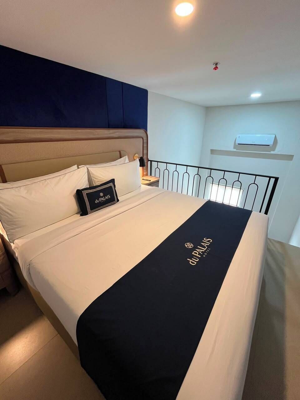 Una habitación de hotel con una cama, una mesita de noche, un aire acondicionado de pared y una lámpara en el techo.