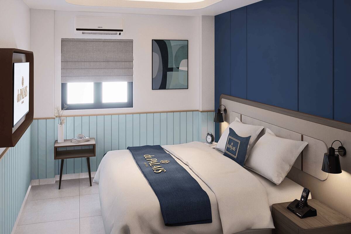 A hotel room with a bed, bedside table, lamp, and a wall-mounted air conditioner.