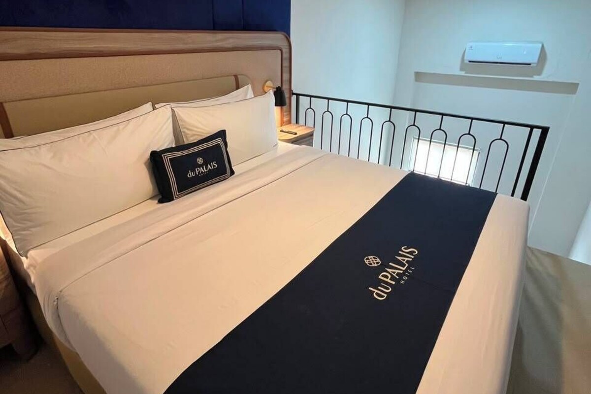A hotel room with a bed, a nightstand, a wall-mounted air conditioner, and a ceiling light.