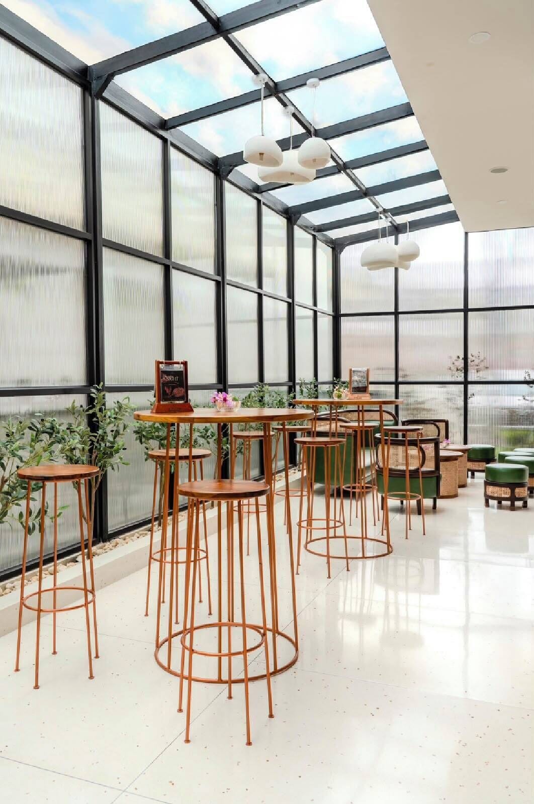 A spacious indoor area with a high ceiling and large windows, featuring multiple tables and chairs arranged for seating.