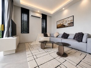 Ein modernes Wohnzimmer mit einer Couch, zwei Couchtischen, einem Flachbildfernseher auf einem Standfuß, einer Klimaanlage und einem Bild an der Wand.
