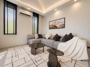 A modern living room with a grey sofa, two glass-top side tables, a white throw blanket, and a painting on the wall. - Villa Luna 2BR Private Plunge Pool (Si Sunthon)