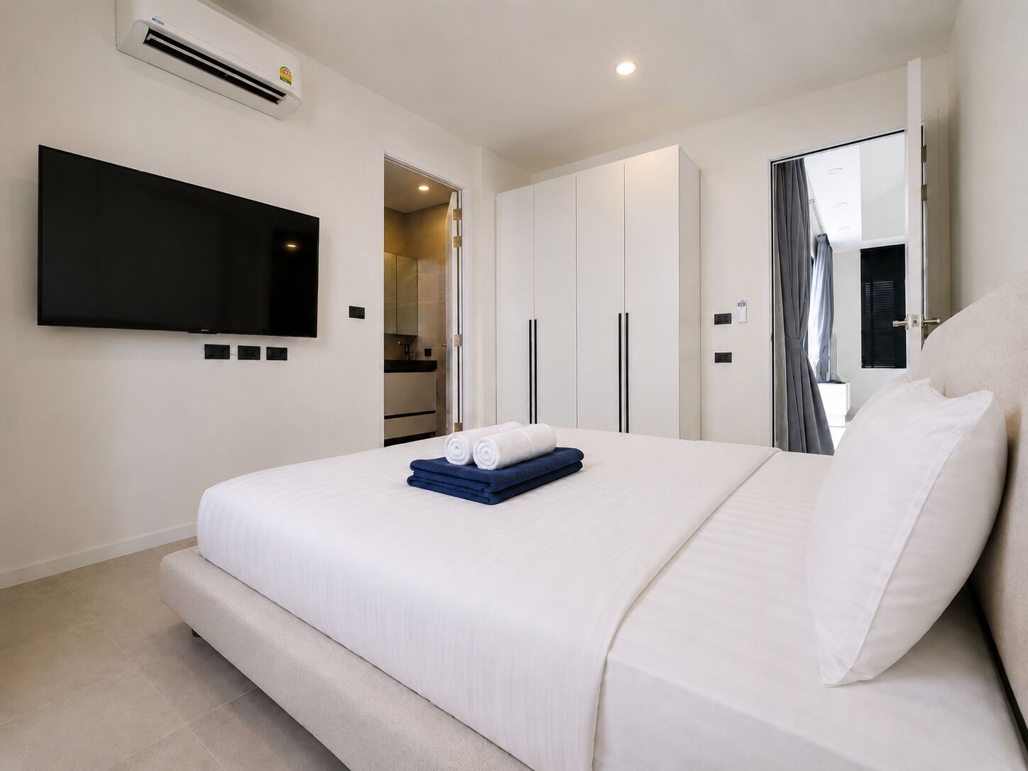 Quarto de hotel moderno com televisão de tela plana, cama grande com roupa de cama branca, closet espaçoso e banheiro visível através de uma porta aberta.