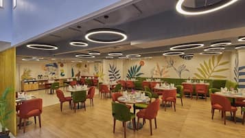 A restaurant with round tables and chairs, a ceiling with circular light fixtures, and a mural with plant designs on the wall.