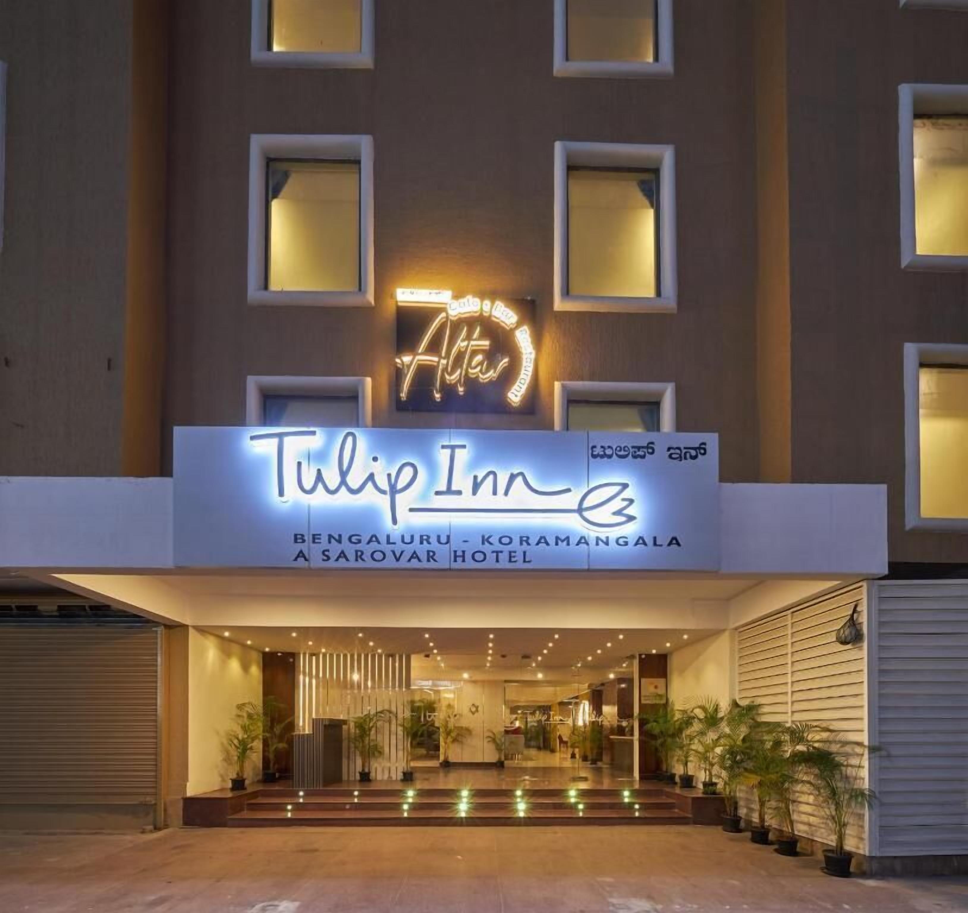 The exterior of Tulip Inn, featuring a neon sign, a modern entrance with potted plants, and a well-lit facade.