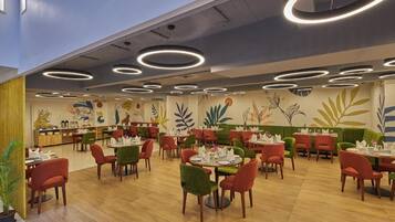 A restaurant with round tables and chairs, a ceiling with circular light fixtures, and a mural with plant designs on the wall.