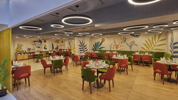 Un restaurante con mesas y sillas redondas, un techo con luminarias circulares y un mural con motivos vegetales en la pared.