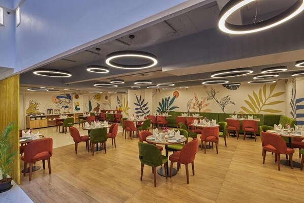 Un restaurante con mesas y sillas redondas, un techo con luminarias circulares y un mural con motivos vegetales en la pared.