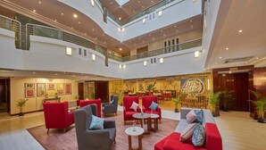 Um saguão de hotel em vários níveis, com assentos em vermelho e cinza, uma área de jantar central e um grande letreiro com a inscrição 'Alar'.