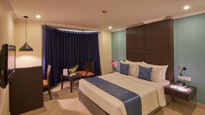 Quarto de hotel com cama, escrivaninha, cadeira, televisão e mesa de cabeceira.
