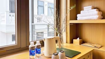 Habitación de hotel con escritorio de madera, un florero con ramas, una pila de toallas y una ventana con vistas a edificios.