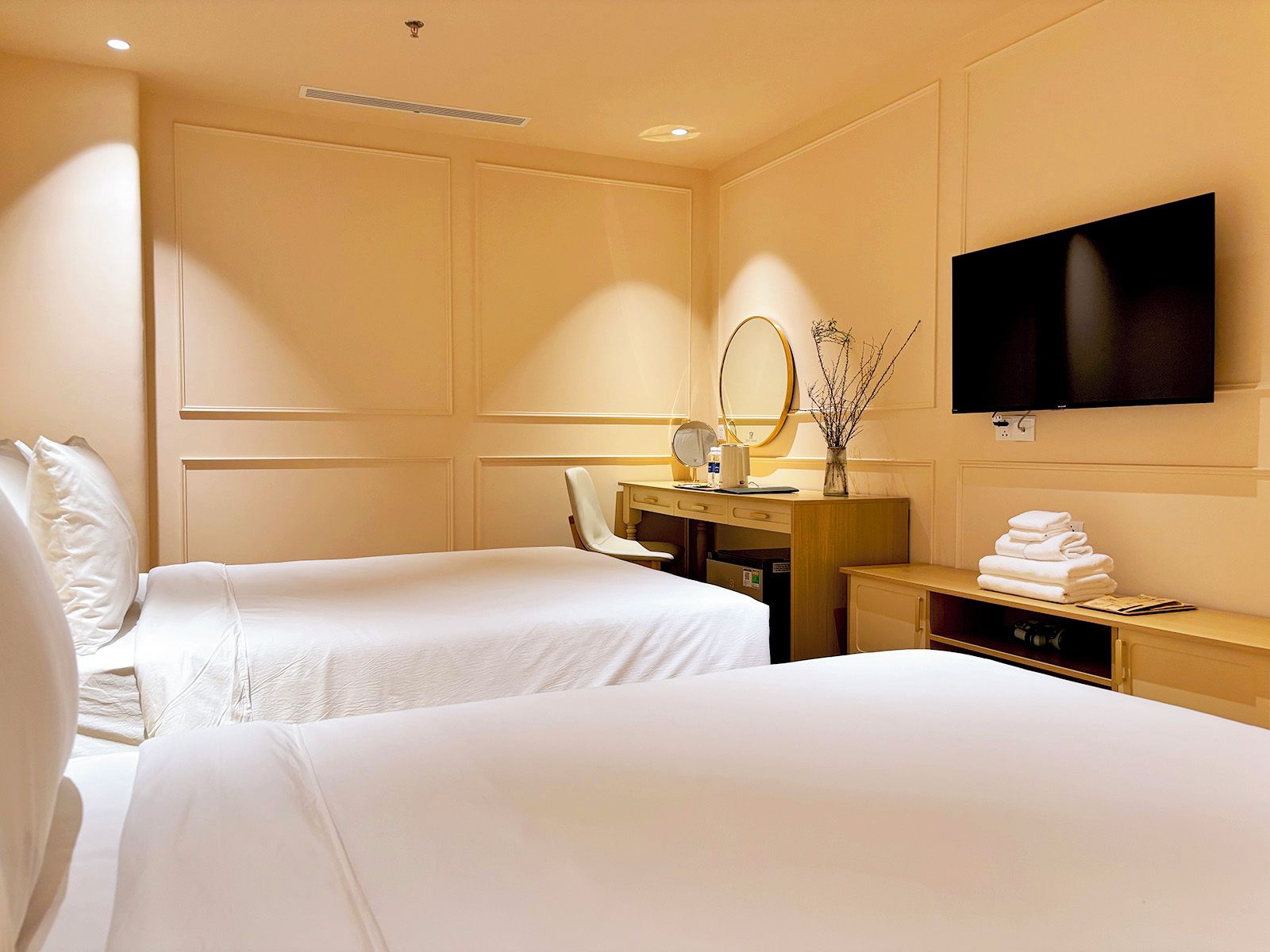 Une chambre d’hôtel avec deux lits, un bureau avec un miroir, une télévision et un vase décoré de branches.