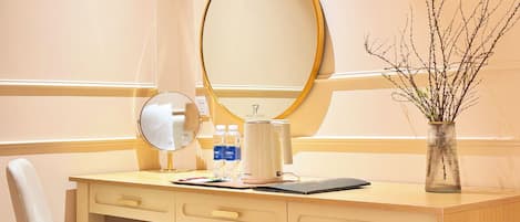 A hotel room with a wooden desk, a mirror, a vase with branches, a chair, and a small trash bin.