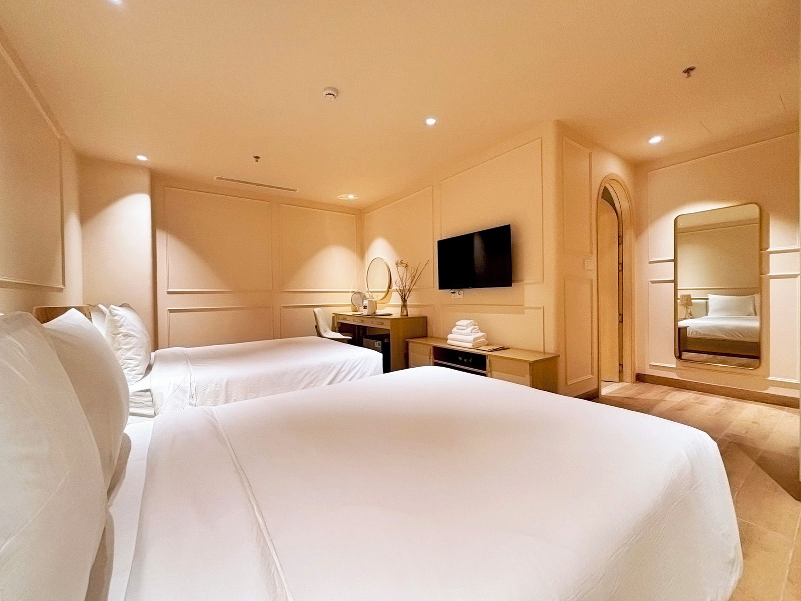 A hotel room with two beds, a desk, a television, a mirror, and a bathroom mirror.