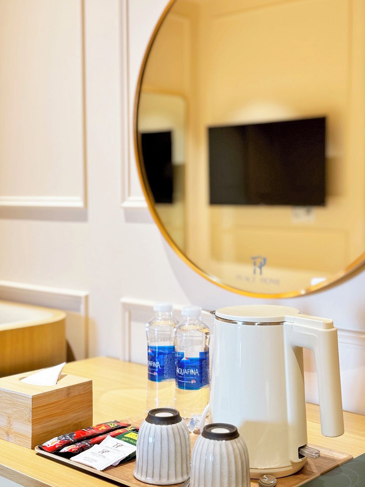 A hotel room with a wooden desk, a mirror, a television, bottled water, a coffee maker, and a tissue box.