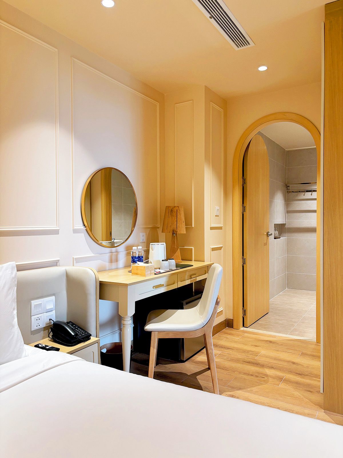 A hotel room with a bed, a desk, a chair, a mirror, a lamp, and a shower area.