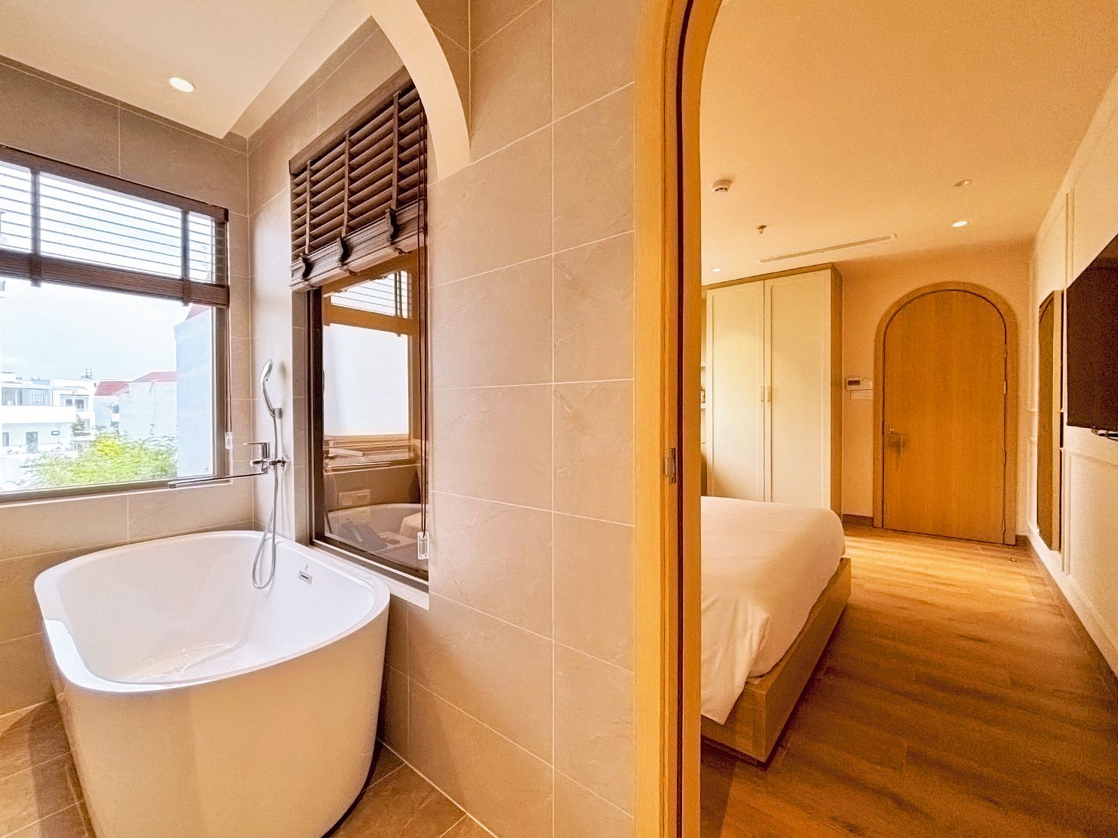 Une salle de bain moderne avec une baignoire sur pieds, une fenêtre avec des stores et une porte en bois donnant sur une chambre.