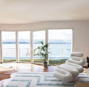 A spacious room with large windows offering a view of the sea and boats, a modern spiral chair, and a potted plant.