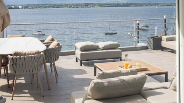 A terrace with a table, chairs, and a sofa, overlooking a body of water with boats and mountains in the distance.