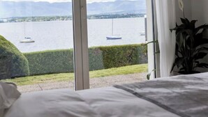 A bedroom with a large window overlooking a lake with boats, a hedge, and a plant.