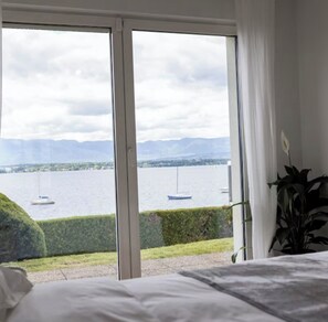 A bedroom with a large window overlooking a lake with boats, a hedge, and a plant.