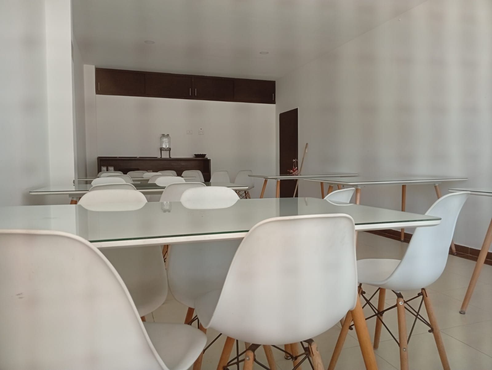 A modern dining area with a glass table and white chairs, a dark wooden cabinet, and a small shelf with a jar and a bowl.