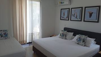 Quarto de hotel com uma cama grande, uma cama menor, uma janela com cortinas translúcidas e três quadros na parede.