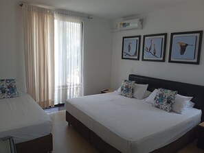 Een hotelkamer met een groot bed, een kleiner bed, een raam met dunne gordijnen en drie ingelijste foto’s aan de muur.