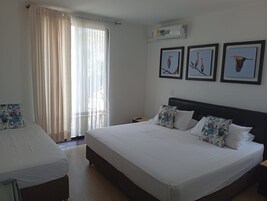 Una habitación de hotel con una cama grande, una cama más pequeña, un ventanal con cortinas traslúcidas y tres cuadros en la pared.