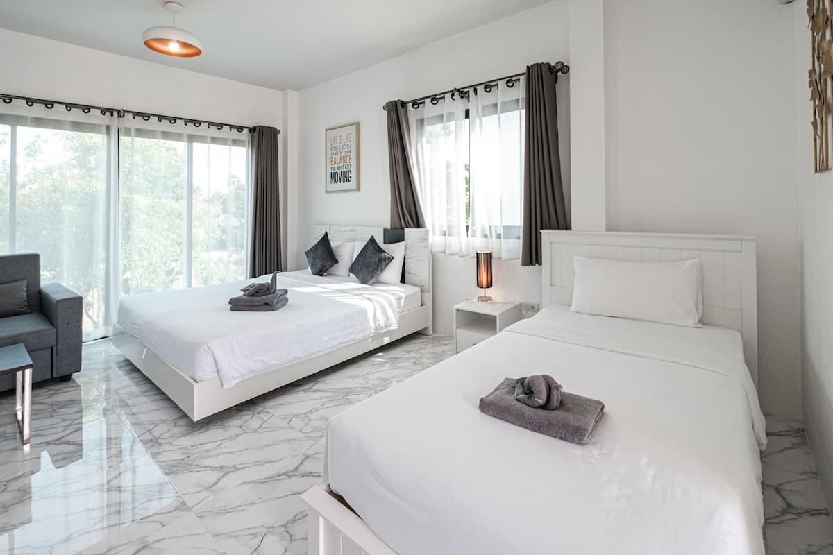 A hotel room with two beds, a sofa, a nightstand, a lamp, and a view of greenery outside.