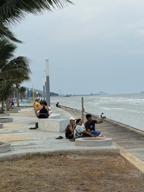 Un passeig marítim amb persones assegudes en bancs de formigó, palmeres i vista al mar.