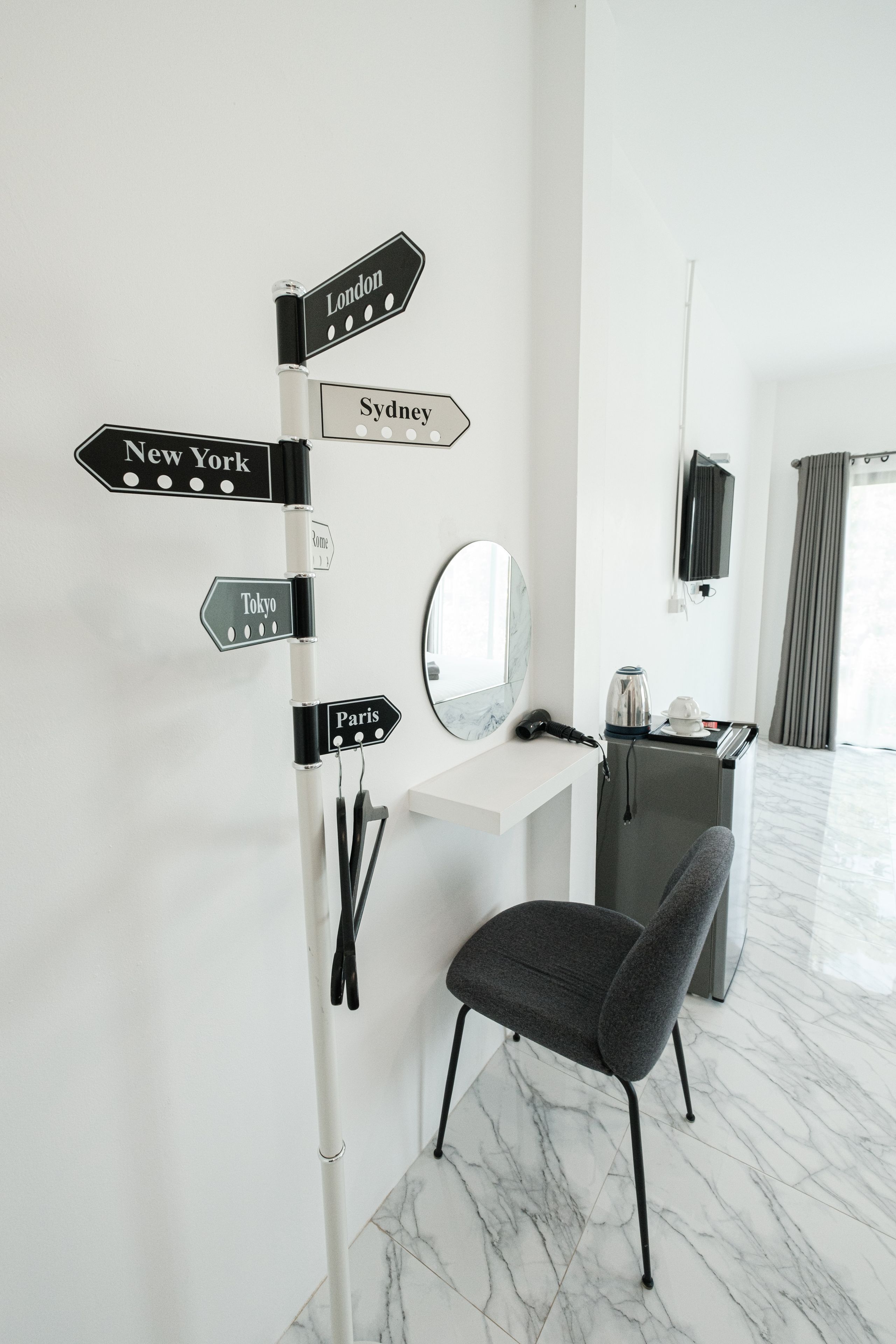 A hotel room with a directional signpost, a mirror, a chair, a desk with a kettle, and a television.