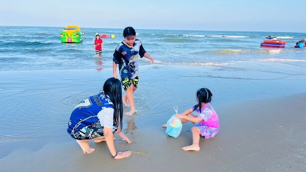 Eine Strandansicht mit spielenden Kindern im Sand, einem grünen aufblasbaren Pool und einem roten Schlauchboot im Hintergrund.