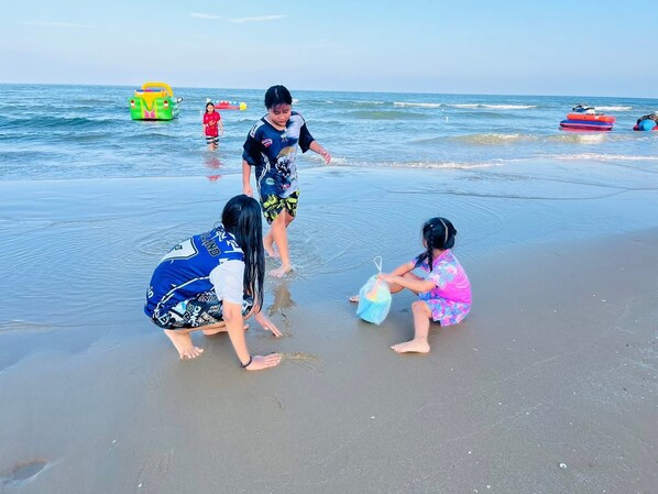 Escena de playa con niños jugando en la arena, una piscina inflable verde y un bote inflable rojo al fondo.