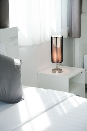 Un dormitori modern amb un llit blanc, una taula de nit amb una làmpada i cortines transparents.