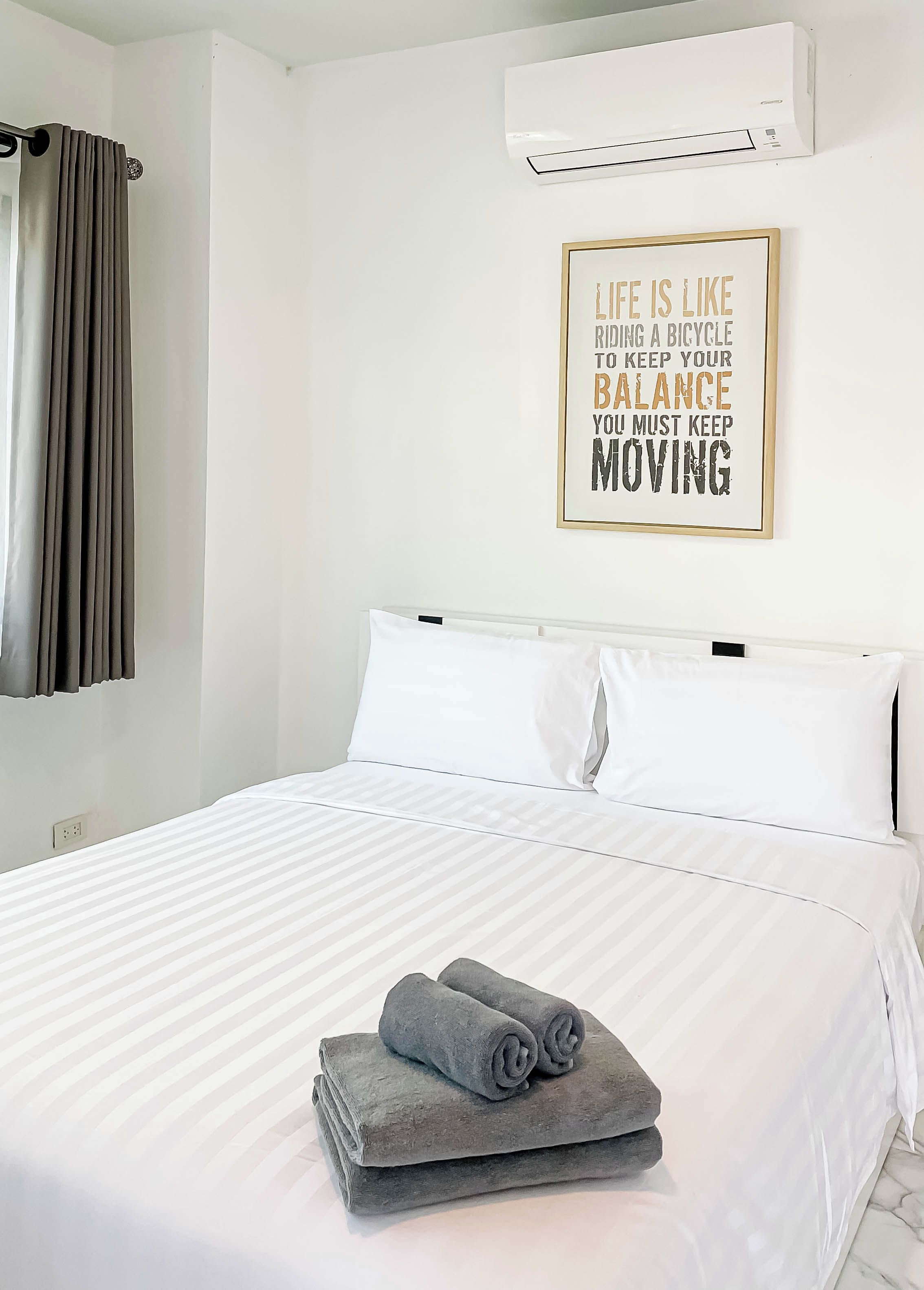 Une chambre d’hôtel avec un lit, un climatiseur fixé au mur, une citation inspirante encadrée et une fenêtre avec des rideaux.