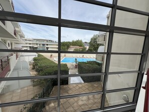 A view through a window of a swimming pool area with a tiled floor, surrounded by greenery and a building with multiple balconies. - Escape to Les Minimes – Pool, Balcony & Private Parking (La Rochelle)