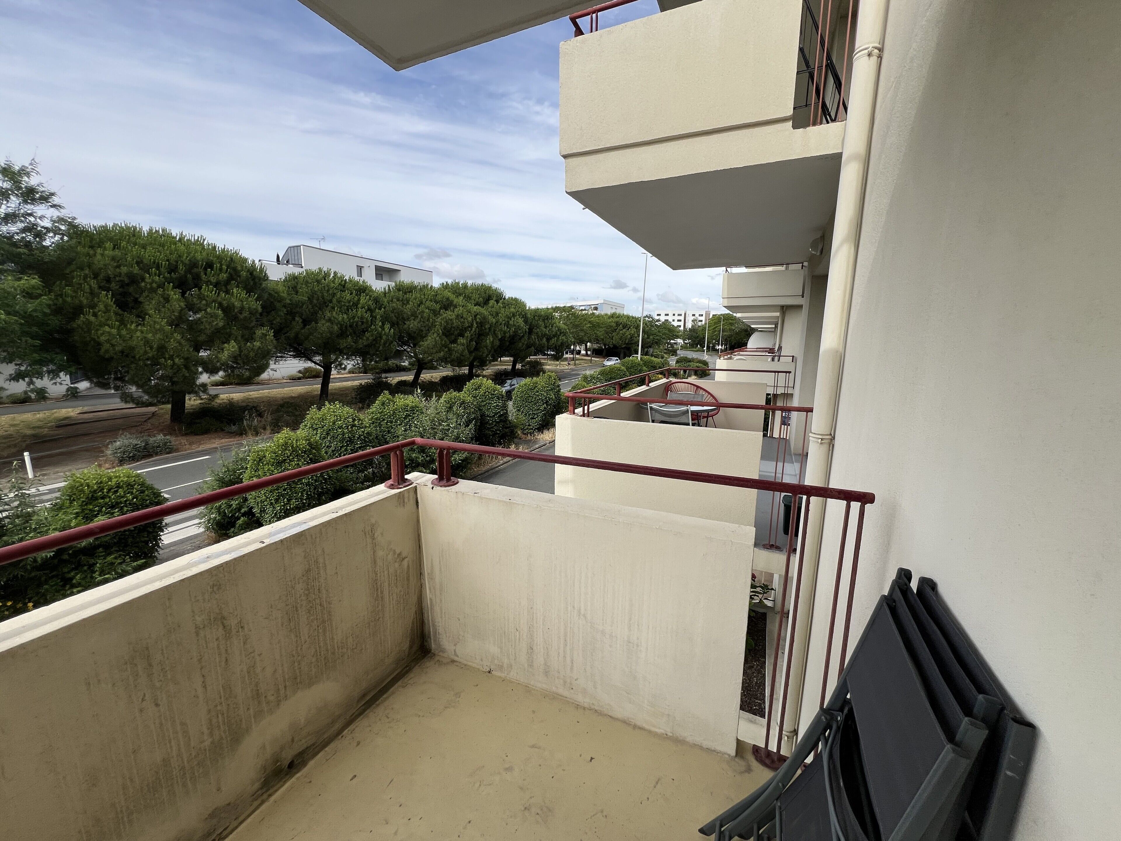A balcony with a view of a road and trees, a red railing, and a building in the background.