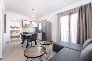 A modern living room with a grey sofa, a dining table with chairs, and a kitchen area with a refrigerator and a hanging light fixture. - Nefeli's Apartment with Sea View and Private Balcony (Kardamaina)